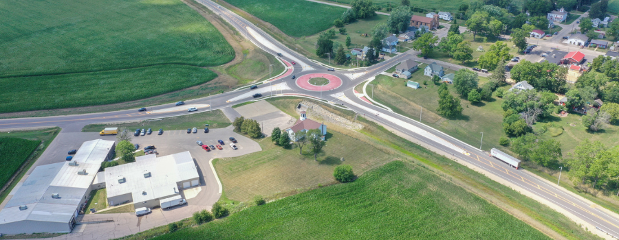 Aerial view of a single-lane roundabout at the intersection of STH 69 and CTH PB, surrounded by lush green farmland and a small residential area.