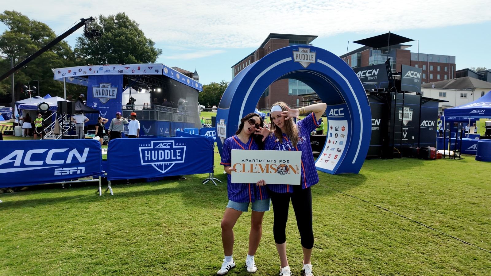 ApartmentsForClemson Interns at the ACC Huddle Two ApartmentsForClemson team members in purple jerseys holding an ApartmentsForClemson sign in front of the ACC Huddle arch at the Stacks