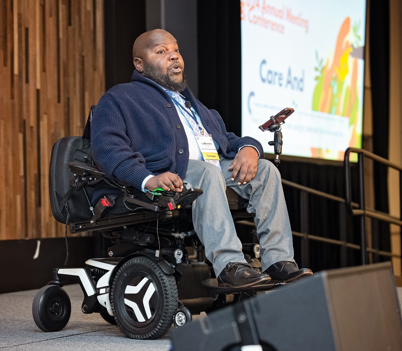 A black man, LaMondre, is in power wheelchair is on stage at a conference. He is wearing a blue sweater, a shirt and tie, and gray slacks. He appears to be in mid-speech during his keynote address..