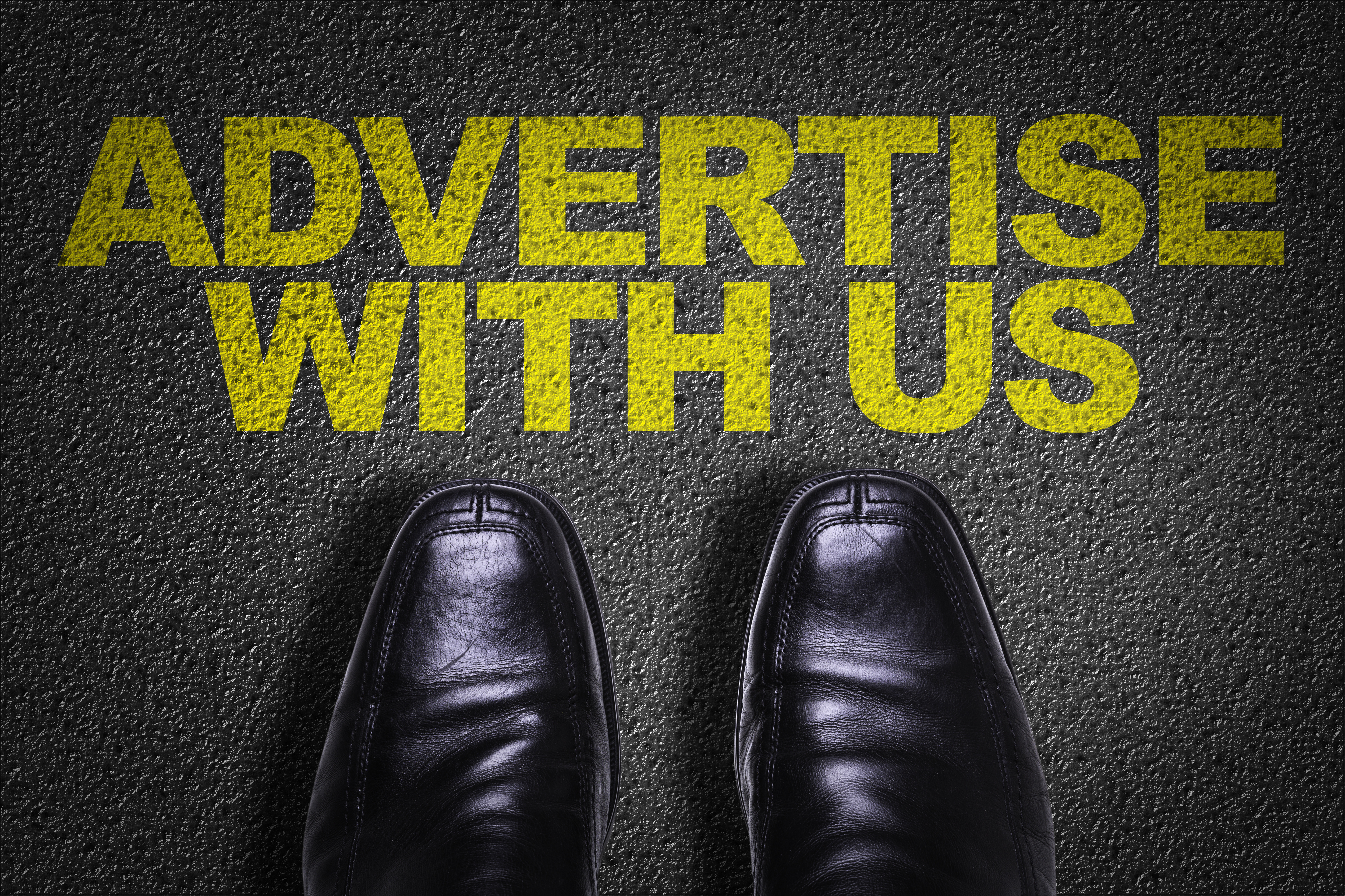 Words 'Advertise with us' painted in yellow on cement with mens black dress shoes standing below them