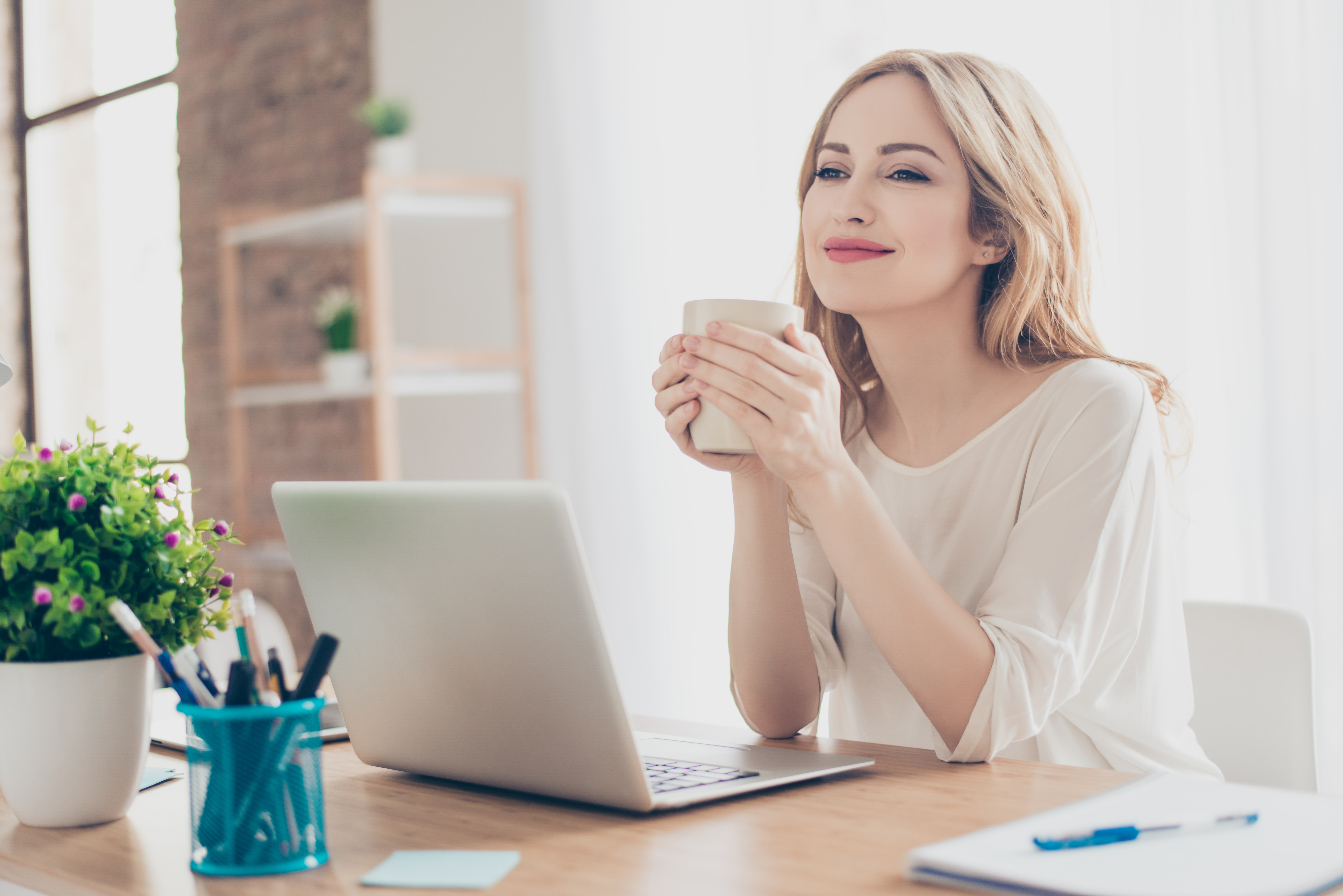 A woman smiles as she sits in front of an open laptop and holds a coffee cup.