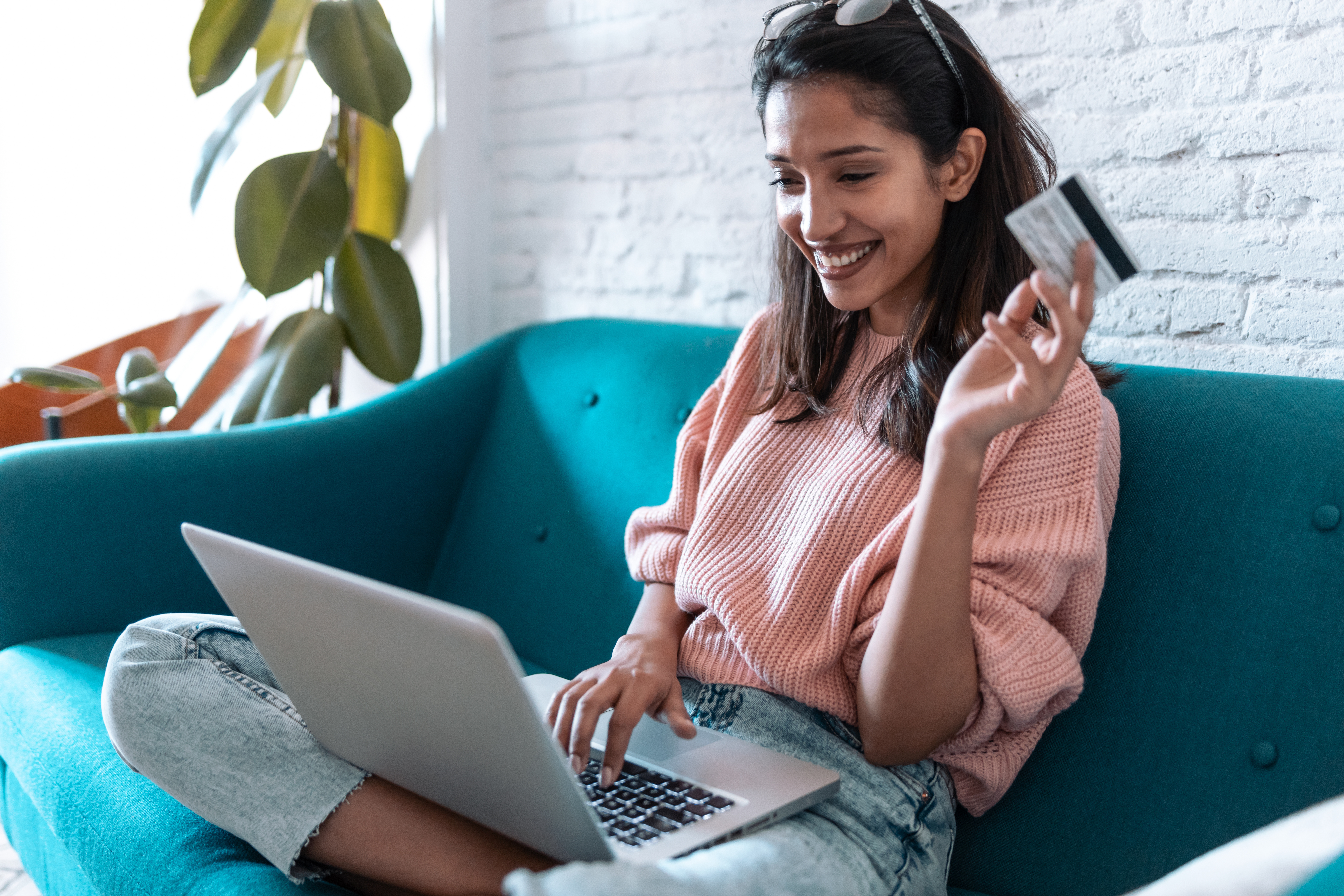 A therapist sits on a blue couch and smiles at an open laptop on her lap. She holds a credit card.