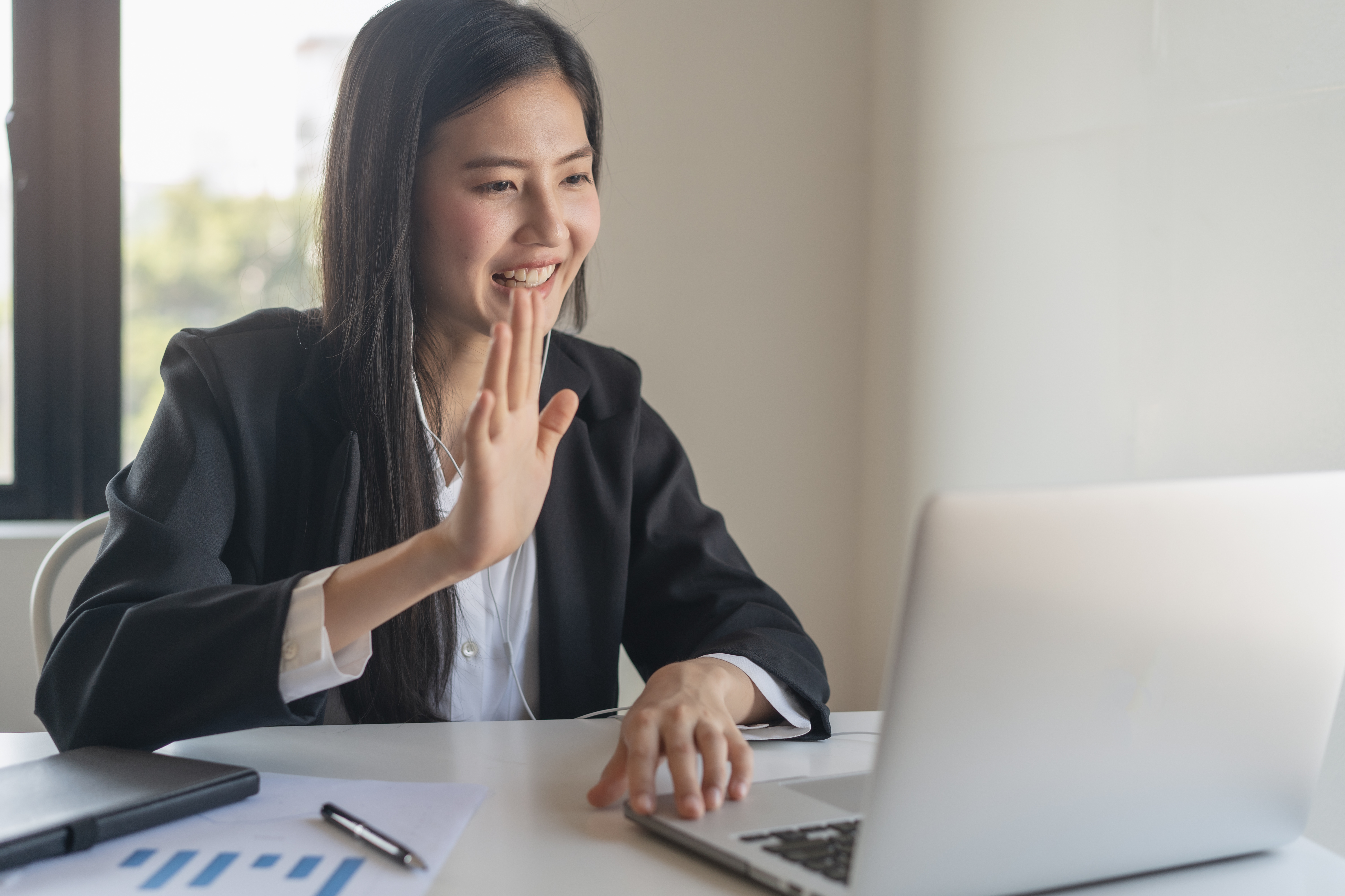 A therapist sits at a desk and smiles and waves at a laptop screen.