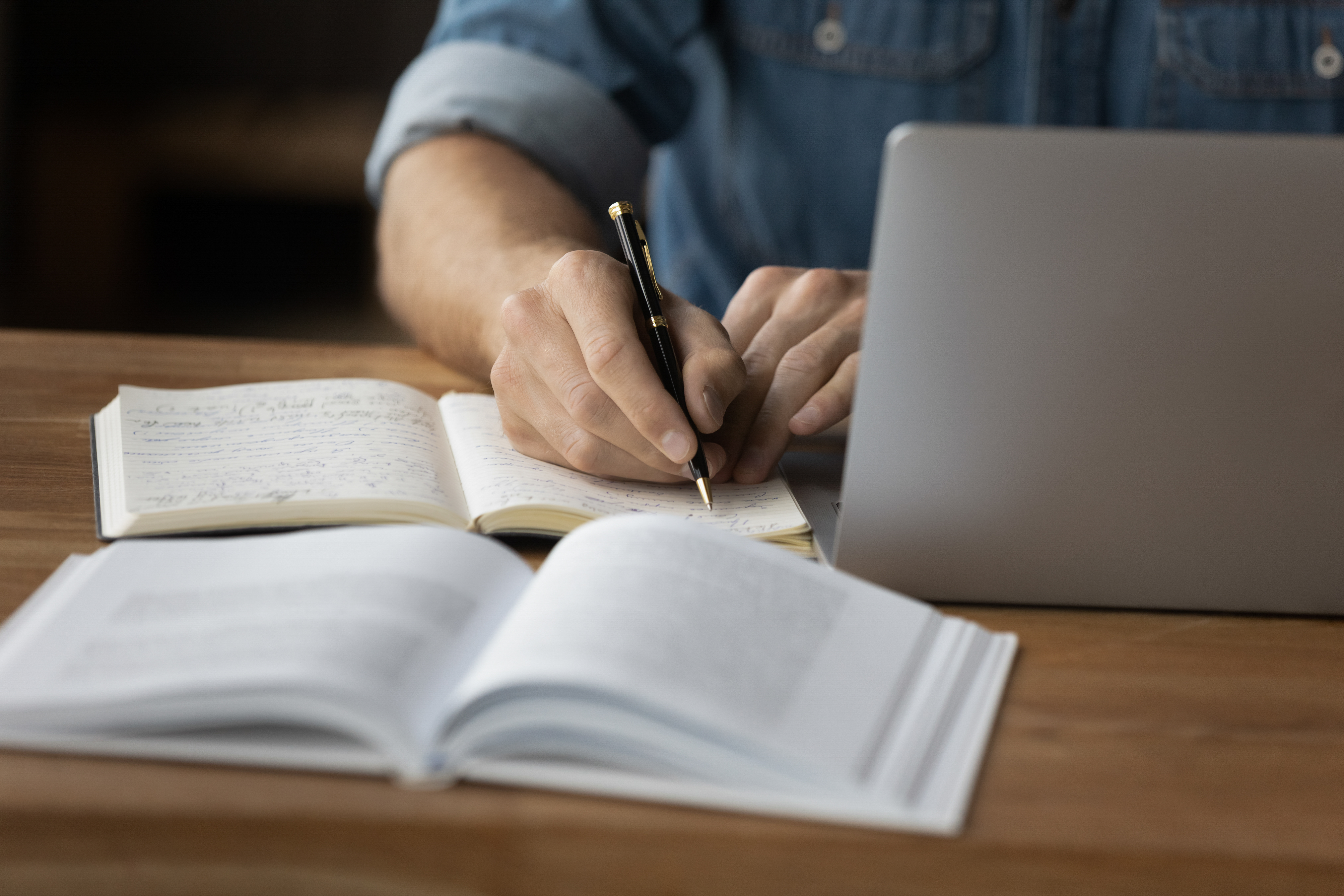 a therapist sits at a laptop and writes in a small notebook, with a textbook open next to them