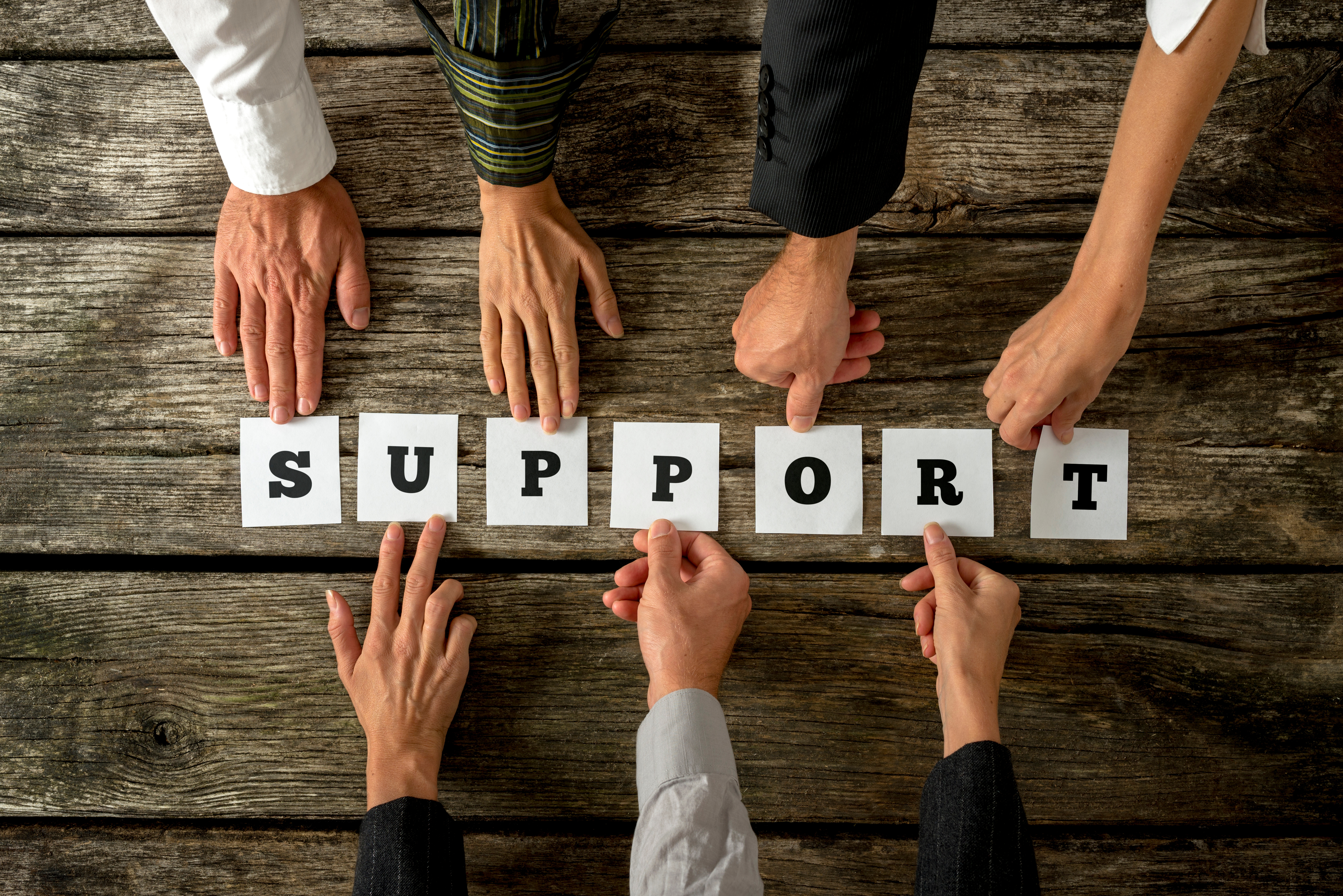 Pictured above: a group of hands lay letters down on a table, spelling "support"