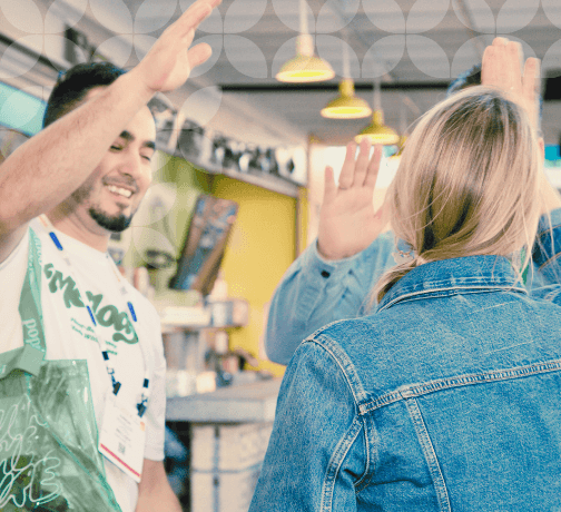 Two people, one wearing a denim jacket, exchange a cheerful high-five in a brightly lit, casual indoor setting, suggesting a moment of camaraderie.