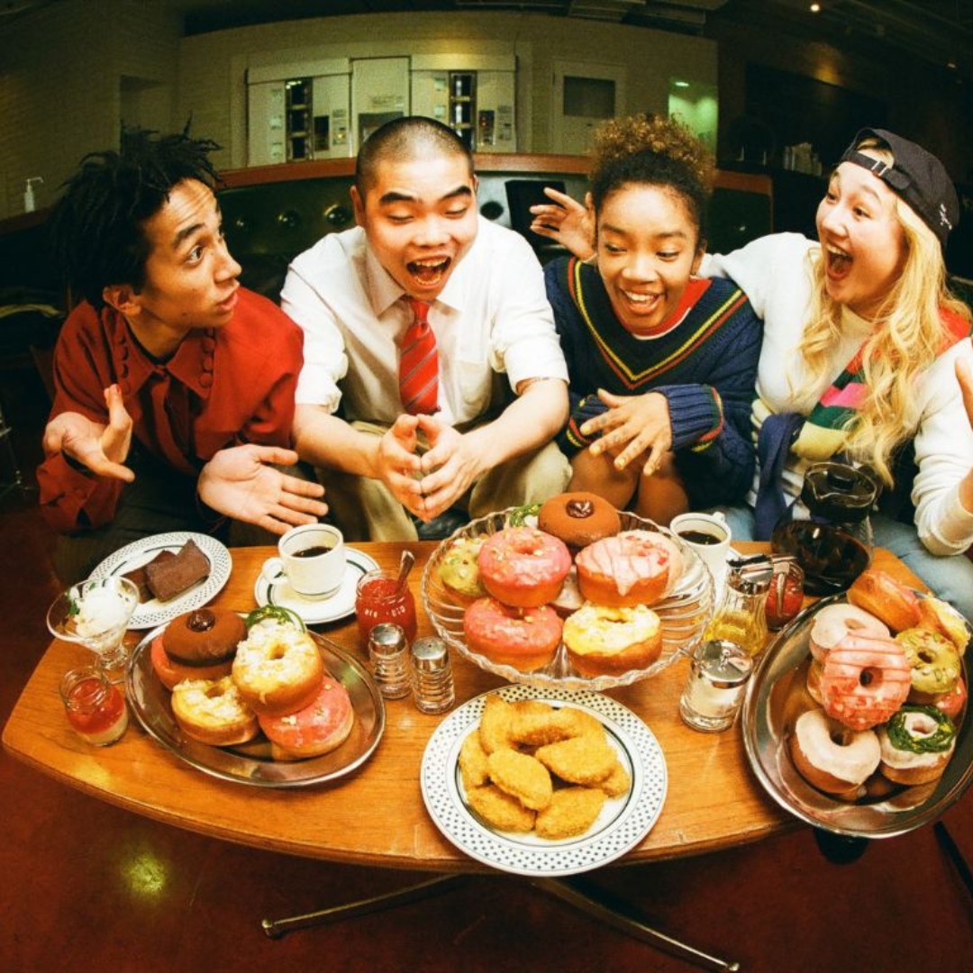 Four friends laugh and react with excitement around a coffee table piled high with colorful frosted donuts, coffee, and pastries. The table features a tiered glass stand in the center holding bright pink, yellow, and chocolate donuts, flanked by additional platters of baked goods and a plate of golden fried food samples.