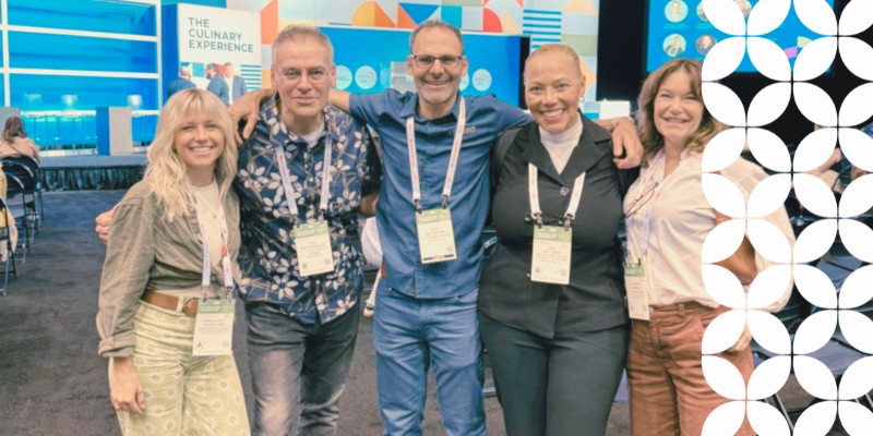 Five smiling professionals stand with their arms around each other at a convention center event. They are wearing event badges on lanyards, and a background sign reads "THE CULINARY EXPERIENCE". A white geometric pattern frames the right border.