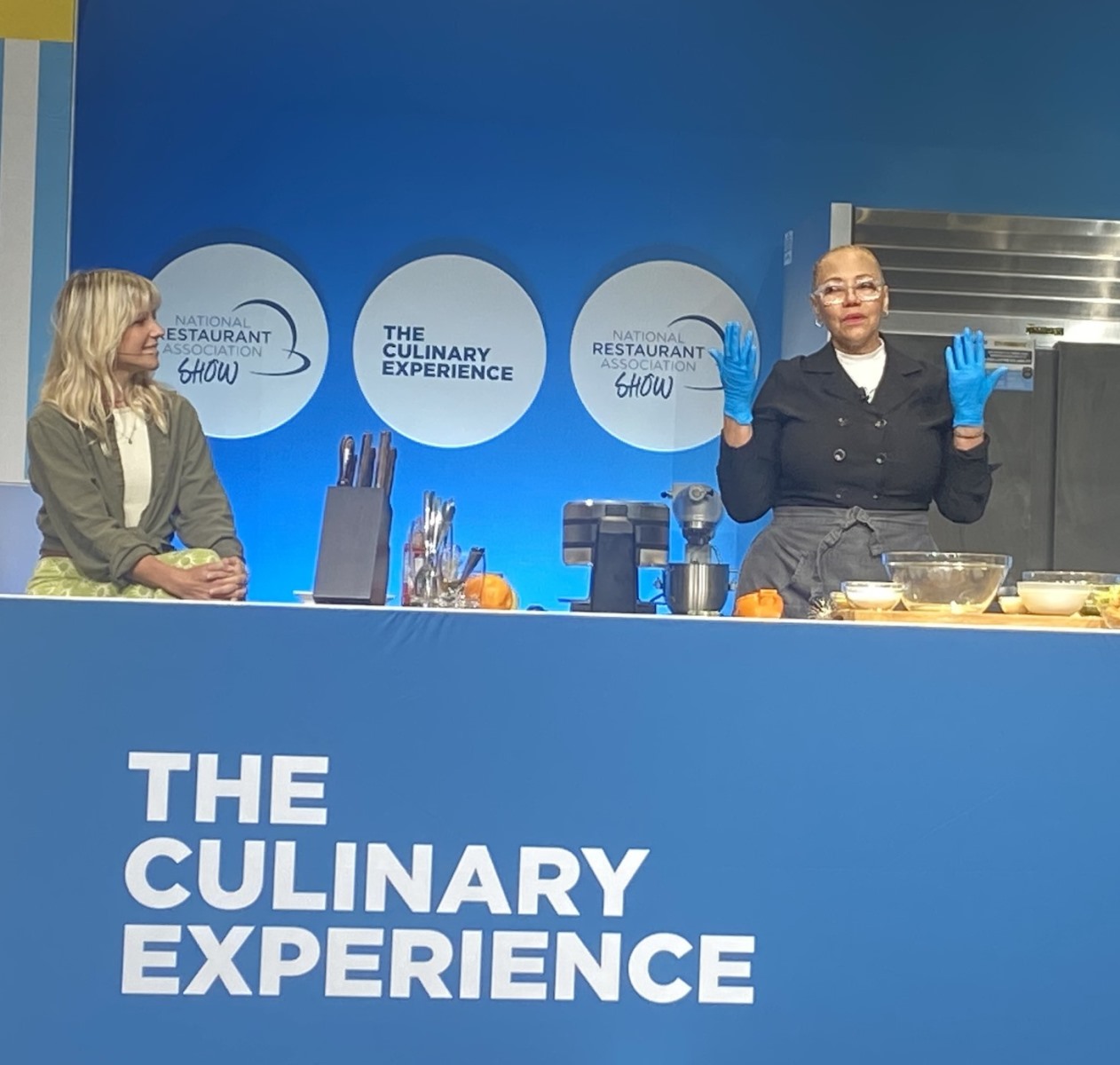 A culinary demonstration stage with a blue background featuring white circle logos for the "National Restaurant Association Show" and "The Culinary Experience". A woman in a chef's coat and blue kitchen gloves gestures with her hands behind a counter containing bowls and a mixer, while another woman sits to the left and watches.