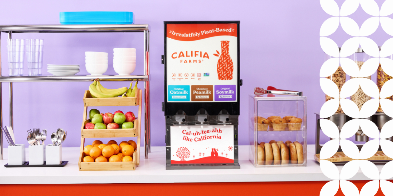 A bright breakfast bar with shelves holding colorful fruits, plates, and cups, next to a Califia Farms plant-based milk dispenser and pastry display, set against a purple wall.