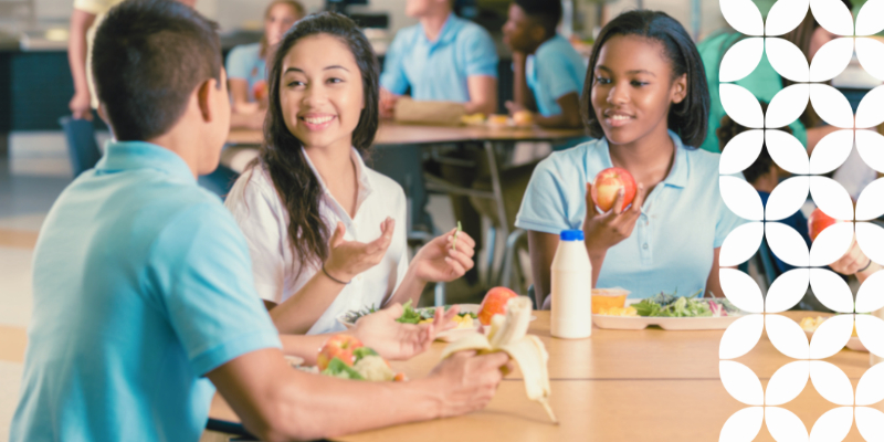 Students sit at a cafeteria table, smiling and talking while eating healthy lunches with fruits and milk. The atmosphere is friendly and lively.
