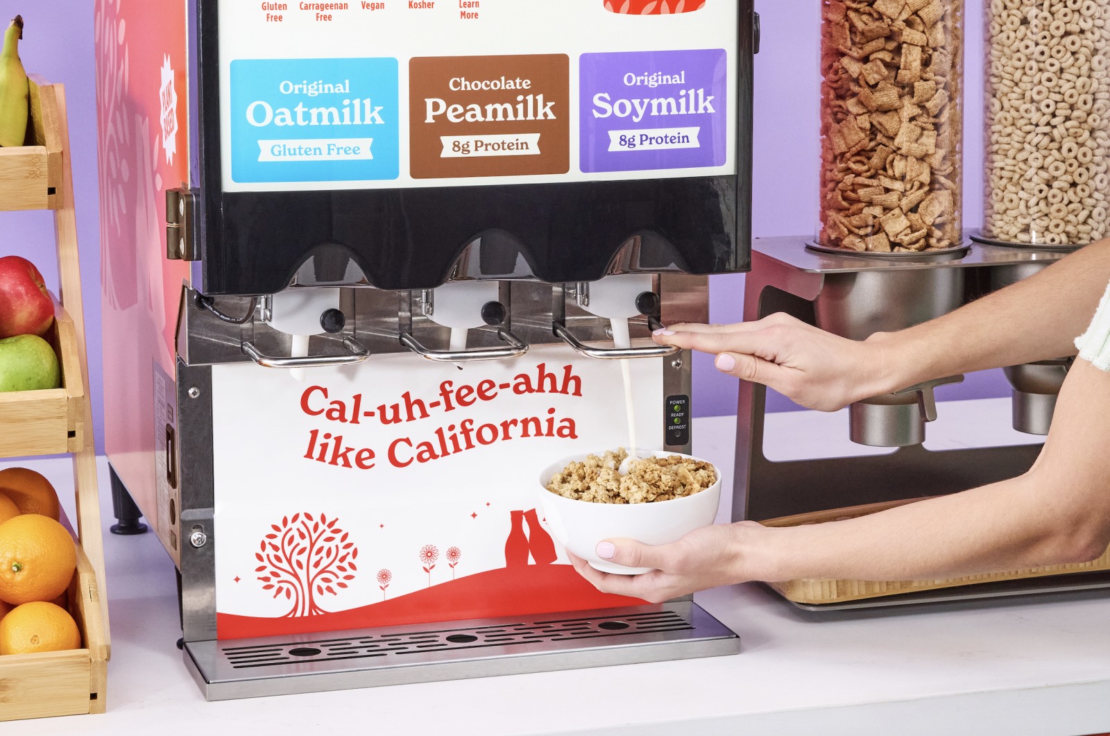Hands dispensing milk into cereal from a machine labeled with oatmilk, peamilk, and soymilk options. Nearby is a cereal dispenser and a fruit basket.