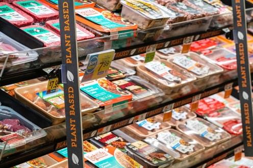 A grocery store refrigerated shelf section filled with packaged plant-based meats, framed by vertical black aisle strips that repeatedly display "PLANT BASED" in bold orange lettering.