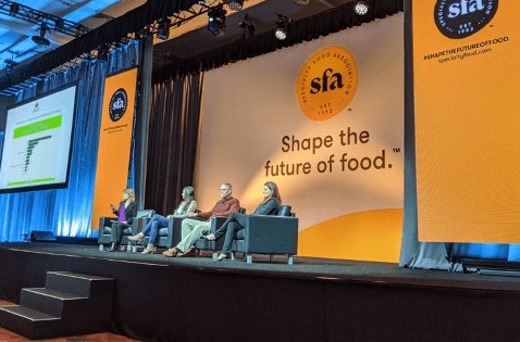 Four speakers sit in armchairs on a stage during a panel presentation at a Specialty Food Association event. A massive backdrop screen behind them features the SFA logo above the tagline "Shape the future of food."