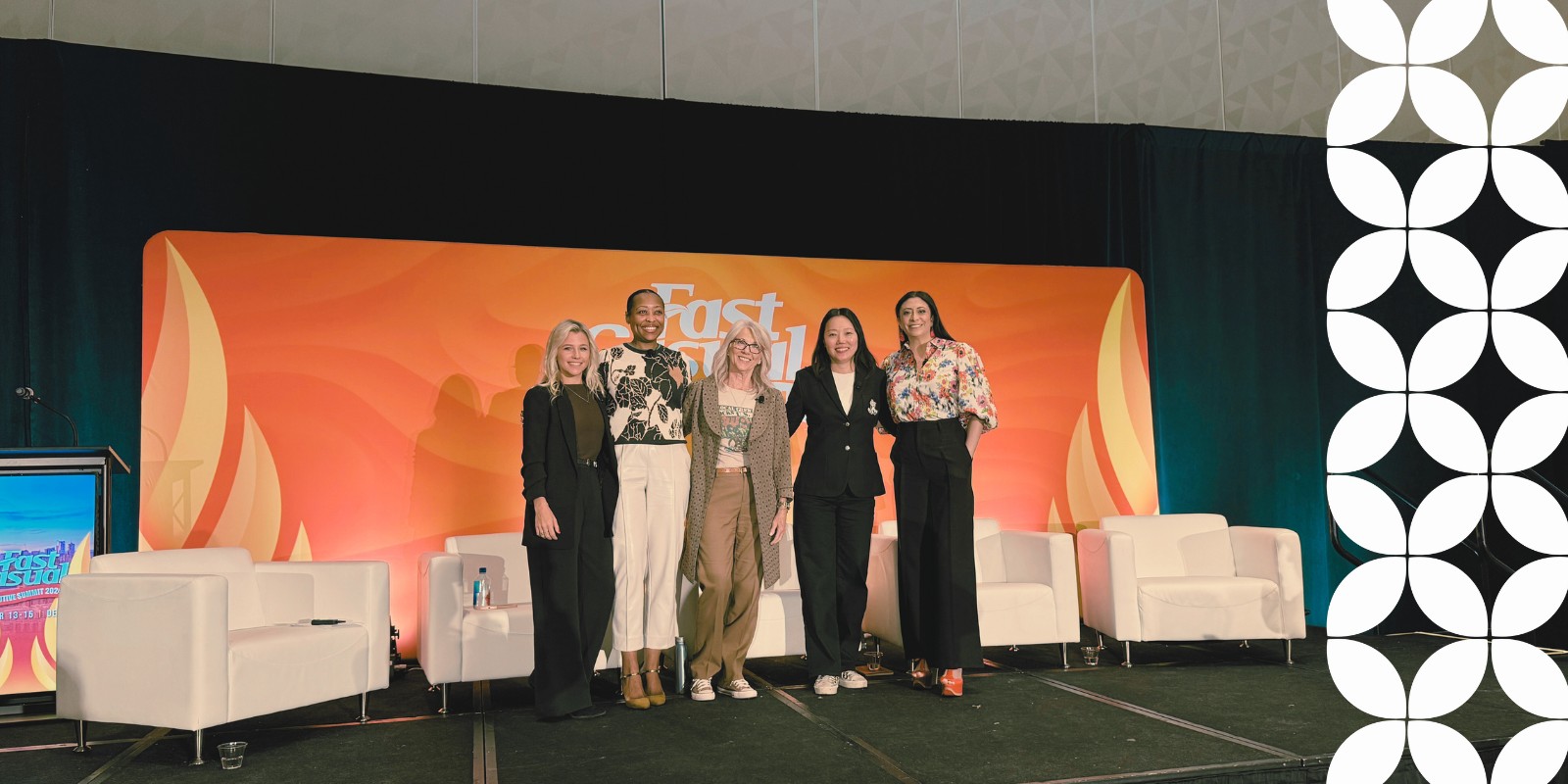 Five smiling women stand shoulder-to-shoulder posing for a group photo on a conference stage. They are positioned in front of an orange background displaying the "Fast Casual" logo, with several empty white armchairs lined up behind them.