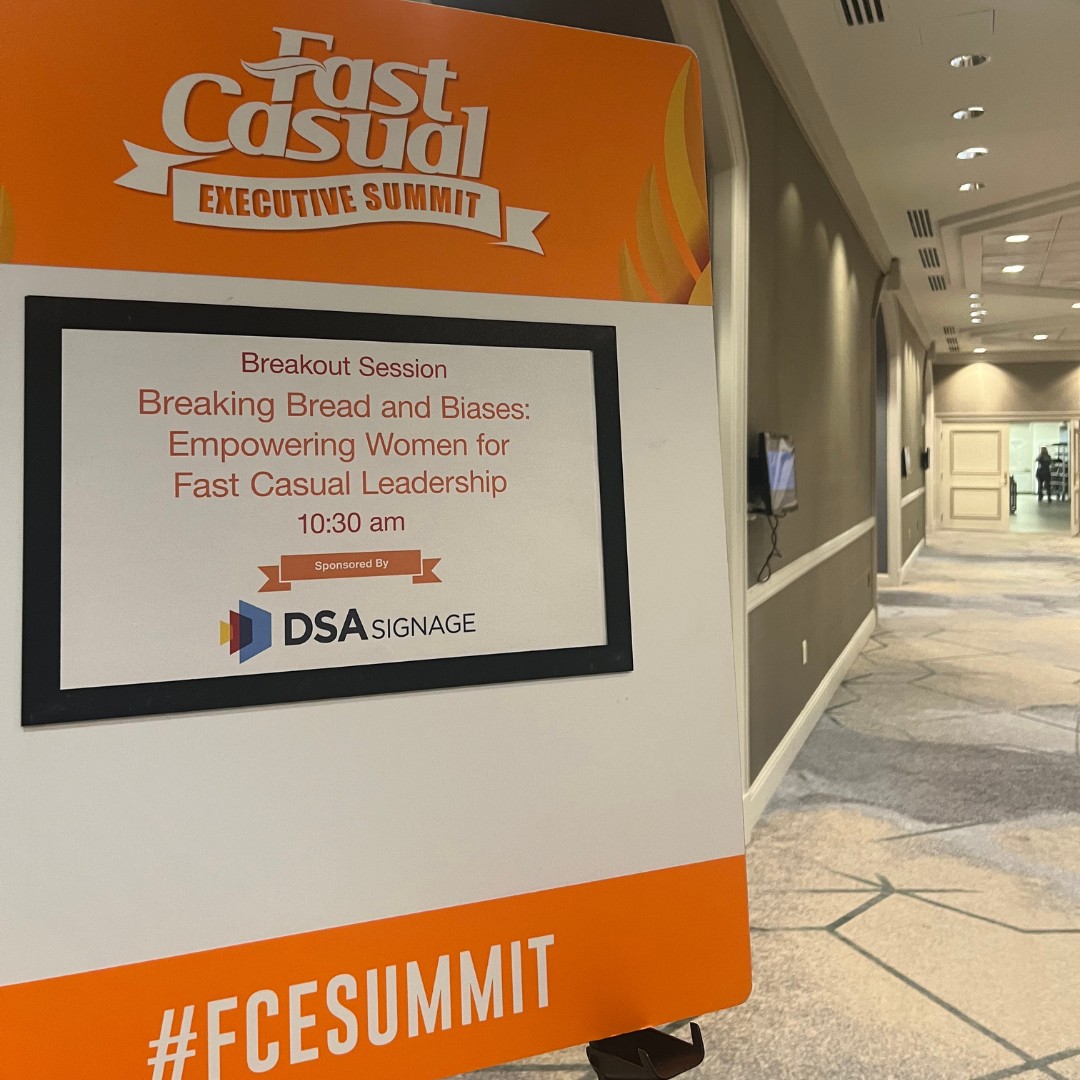 A close-up of an orange and white event sign reading "Fast Casual Executive Summit" and "#FCESUMMIT". The digital screen on the sign displays details for a breakout session titled "Breaking Bread and Biases: Empowering Women for Fast Casual Leadership" at 10:30 am, sponsored by DSA Signage. An empty conference hallway stretches into the background.