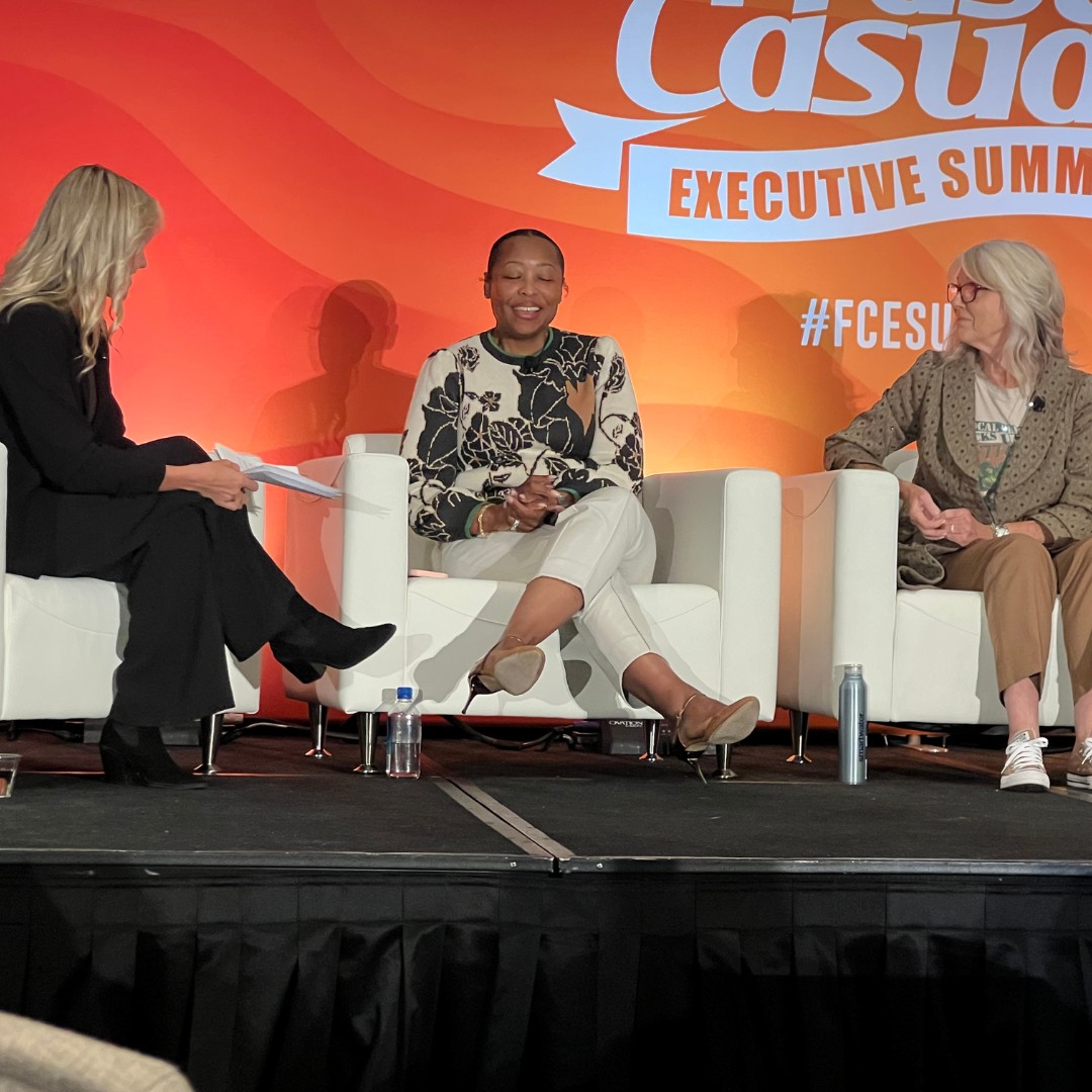 A panel discussion on a stage in front of a vibrant orange backdrop with the "Fast Casual Executive Summit" logo. Three women are seated in white armchairs; the woman on the left in a black suit holds discussion papers, the woman in the center speaks while wearing a floral patterned sweater and white pants, and the woman on the right listens attentively in a patterned jacket and glasses.