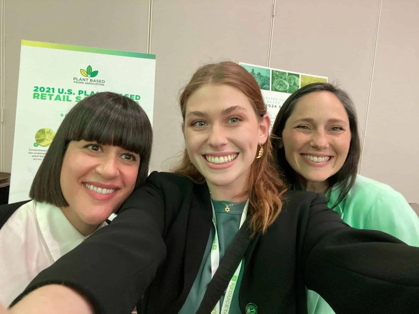 Three smiling women pose for a close-up selfie inside a convention hall. In the background, a large presentation sign from the Plant Based Foods Association features a green infographic titled "2021 U.S. Plant-Based Retail Market".