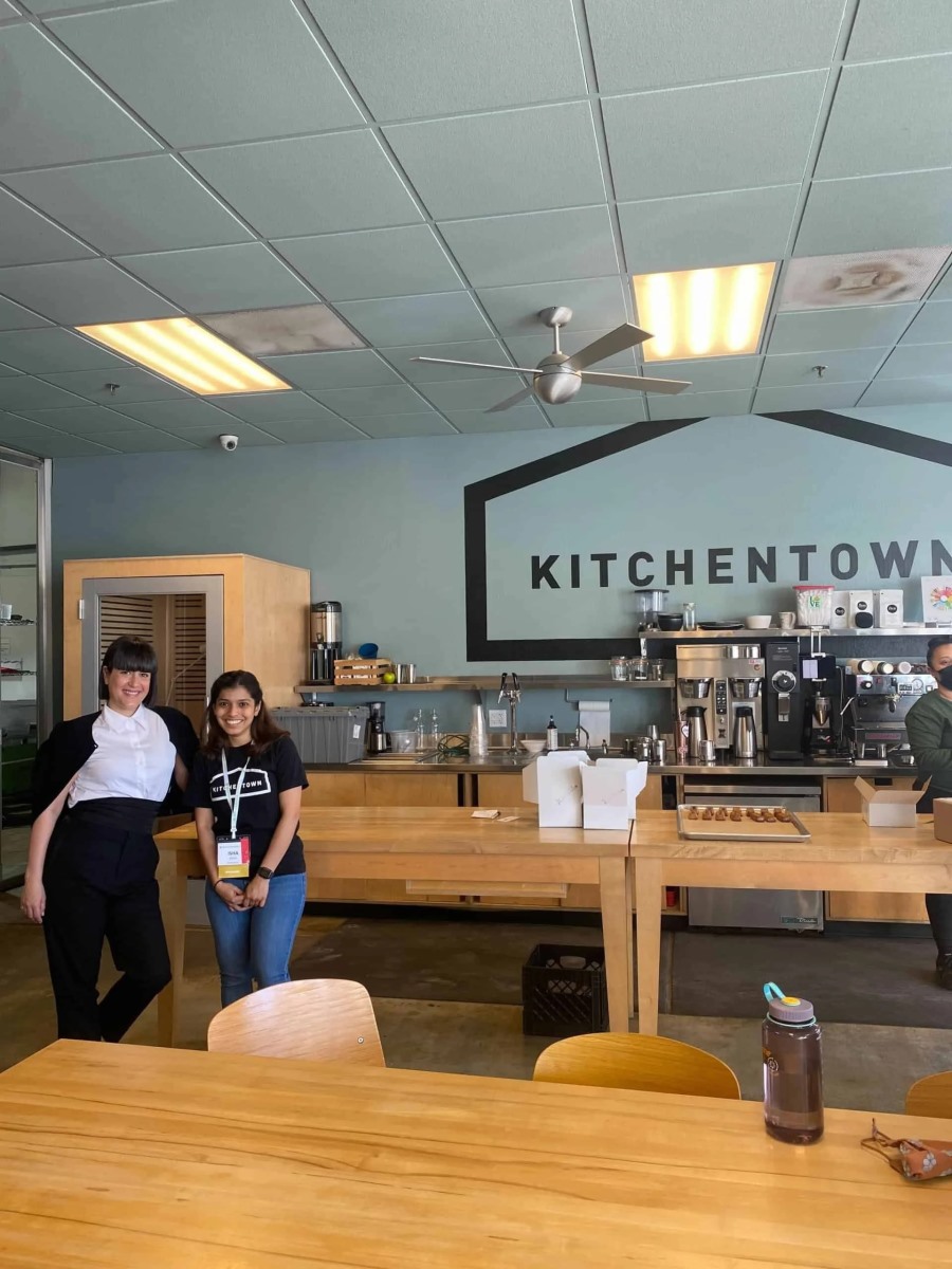 Two women stand together posing next to a long wooden counter inside a commercial kitchen space. A prominent black house outline graphic on the back wall encloses the word "KITCHENTOWN".
