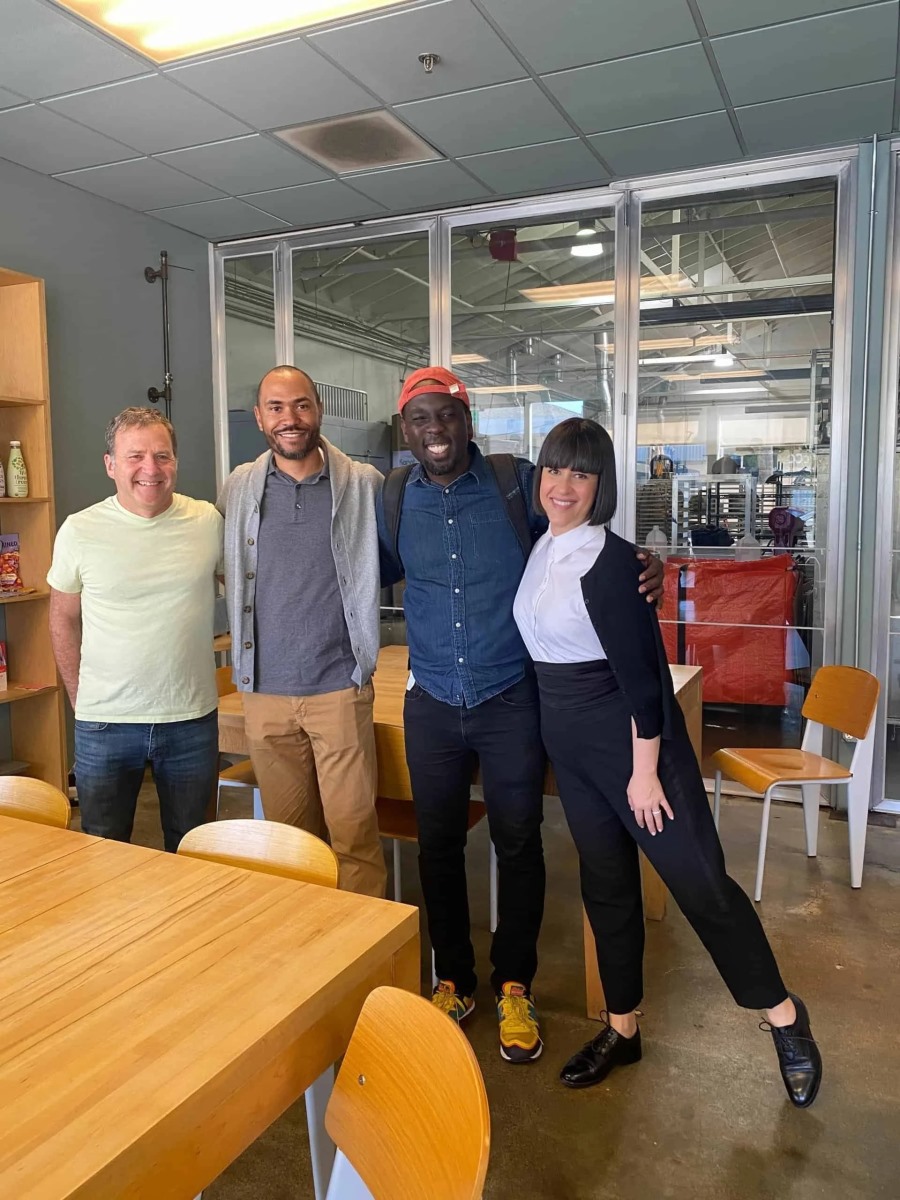 Four professionals stand in a row smiling warmly for a photo in a modern workspace or kitchen area. The group features three men and a woman with straight bangs, posing together next to large glass doors that look out into an industrial production room.