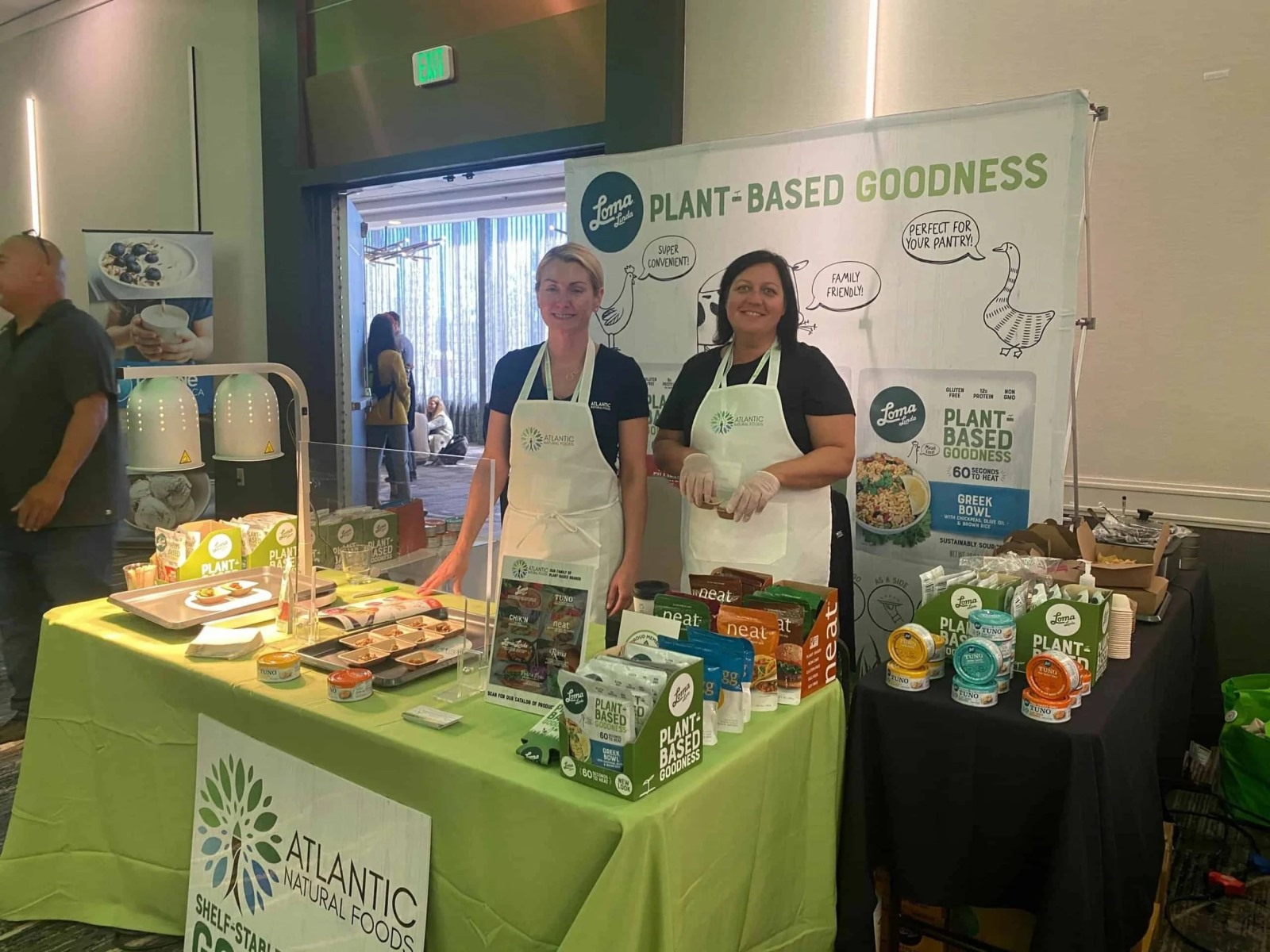 Two women wearing white aprons stand smiling behind a green-clothed exhibition booth promoting Loma Linda "Plant-Based Goodness" and Atlantic Natural Foods. The table is neatly arranged with packaged food bowls, canned goods, and sample trays.