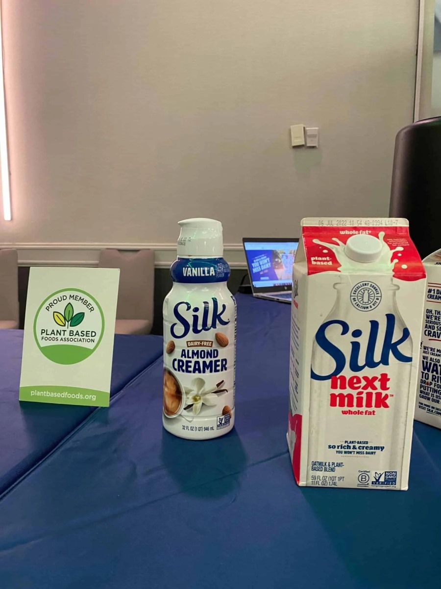 A bottle of Silk Vanilla Almond Creamer and a carton of Silk Next Milk stand side-by-side on a blue table. To the left, a small green table card displays the logo for "PROUD MEMBER PLANT BASED FOODS ASSOCIATION".