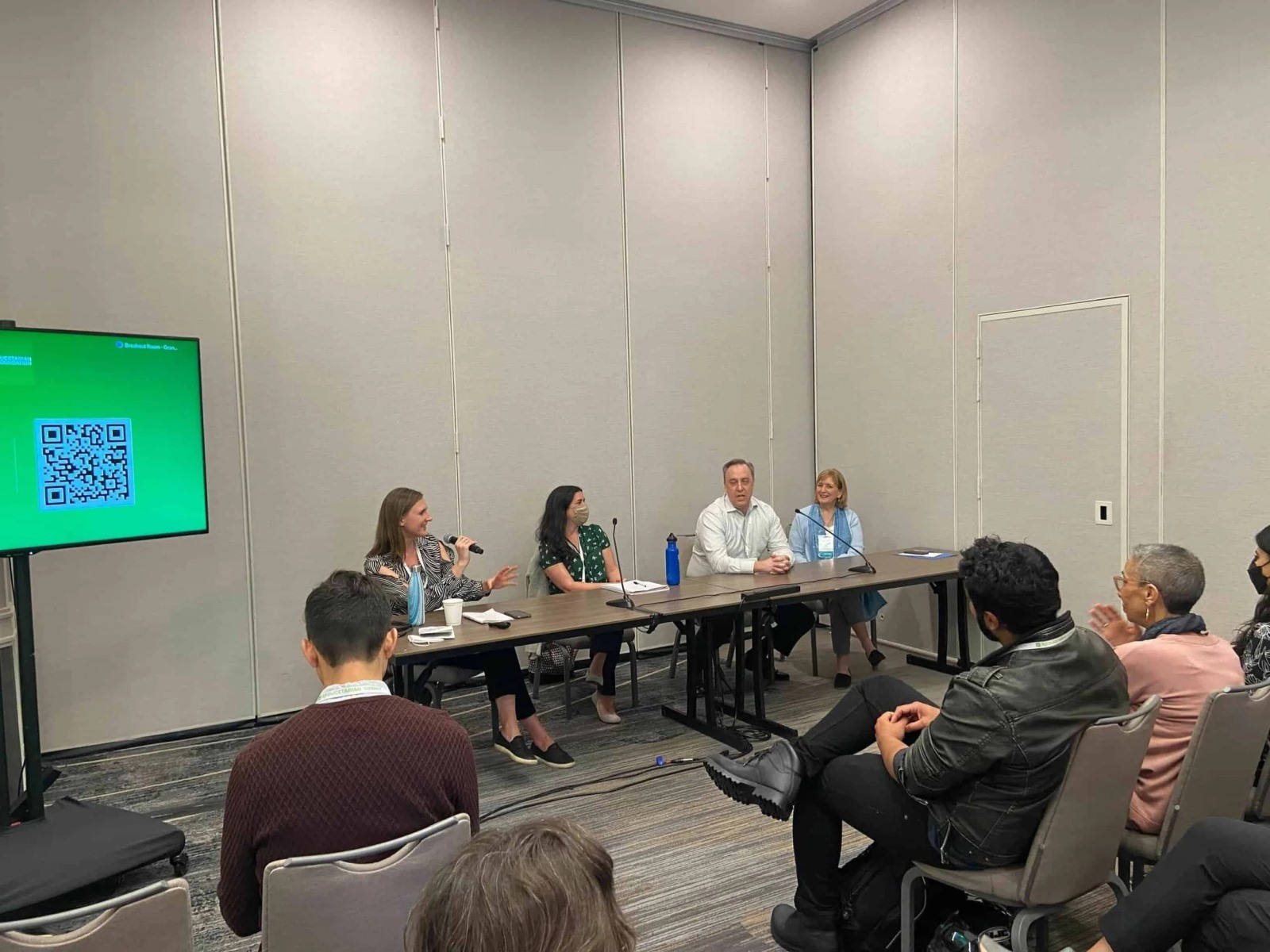A panel of four speakers sits at a long table during a workshop presentation, with one woman speaking into a microphone. Seated audience members listen in the foreground, and a large flat-screen monitor on the left displays a bright green screen with a QR code.