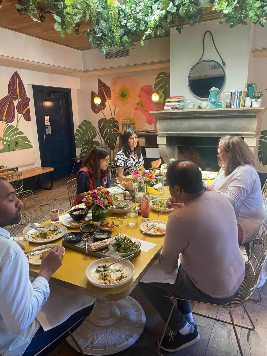 A group of people sharing a meal together around a long yellow dining table inside a vibrant restaurant. The table is filled with assorted small plates, side dishes, and floral centerpieces under a ceiling canopy of hanging green ivy plants.