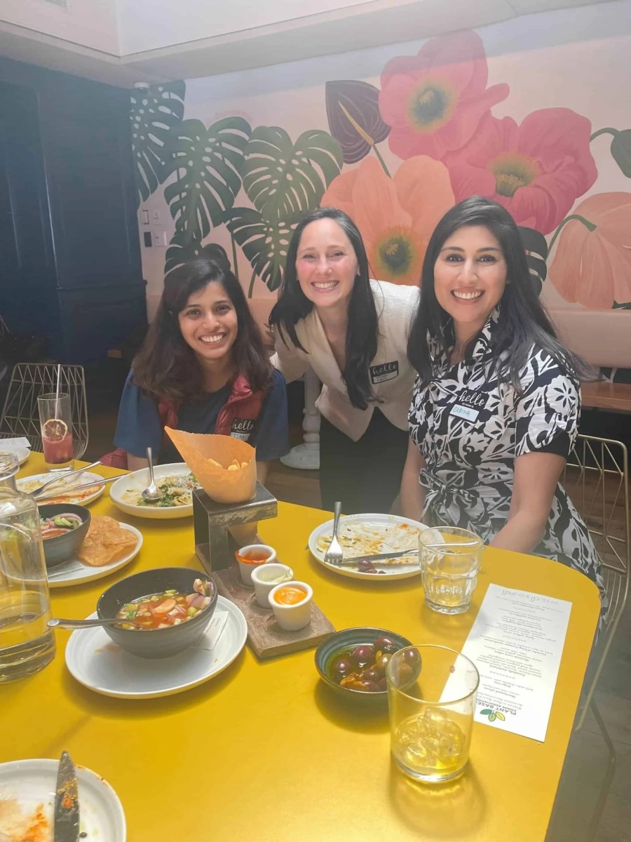 Three women smile warmly at the camera from behind a yellow restaurant table. The table is scattered with small ceramic bowls of appetizers, dips, a basket of chips, and a printed menu.