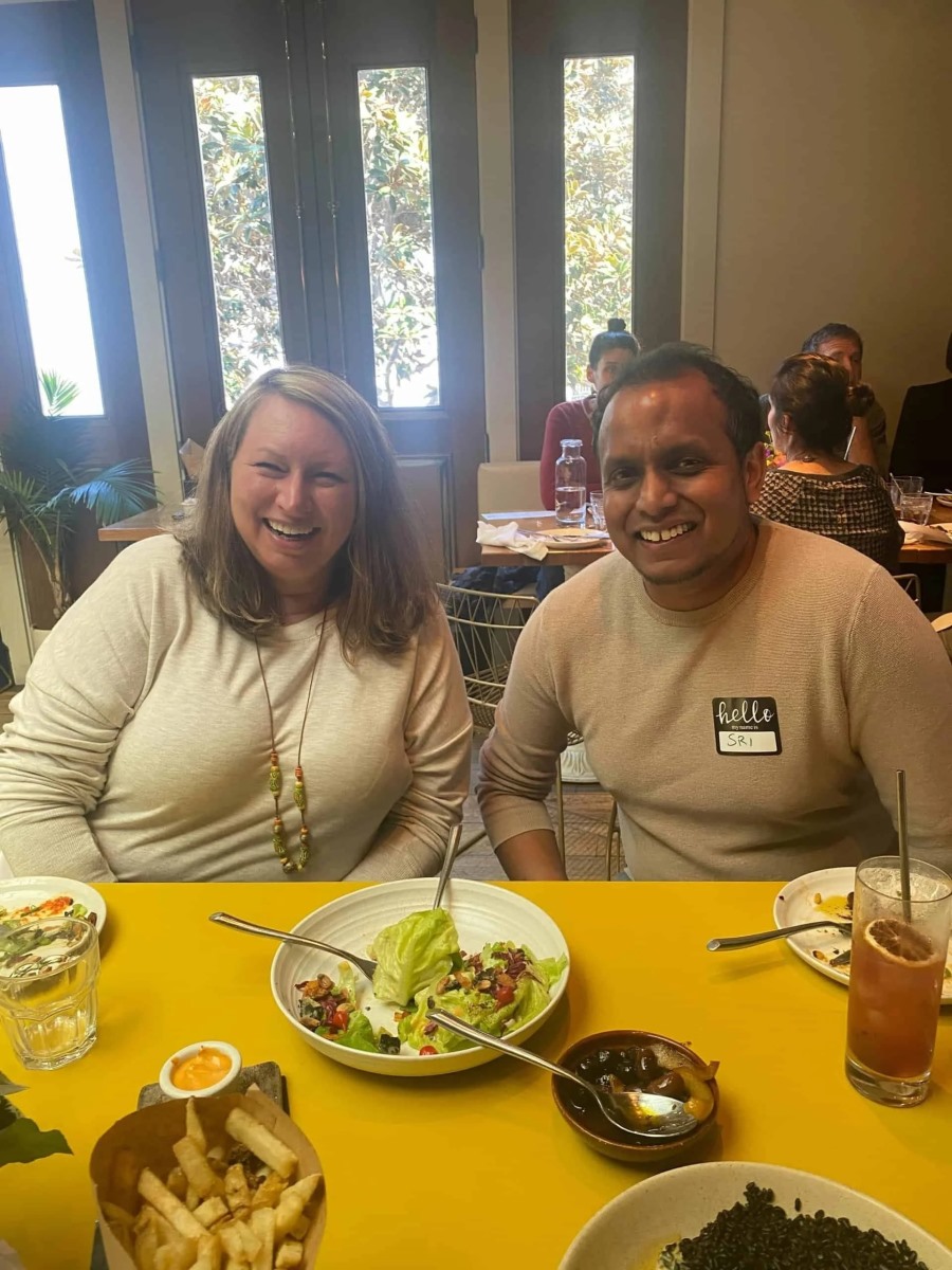 A man wearing a "hello my name is SRI" sticker badge and a woman sit side-by-side laughing at a bright yellow dining table. Plates of green salad, a bowl of olives, a cocktail, and a basket of french fries are spread before them.