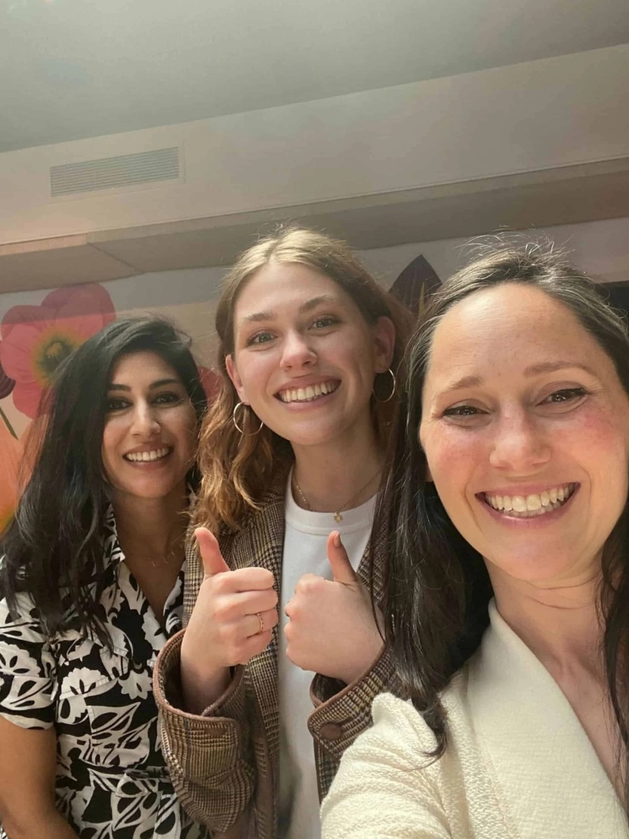 A close-up selfie of three smiling women standing together indoors, with the woman in the center happily giving two thumbs up to the camera.