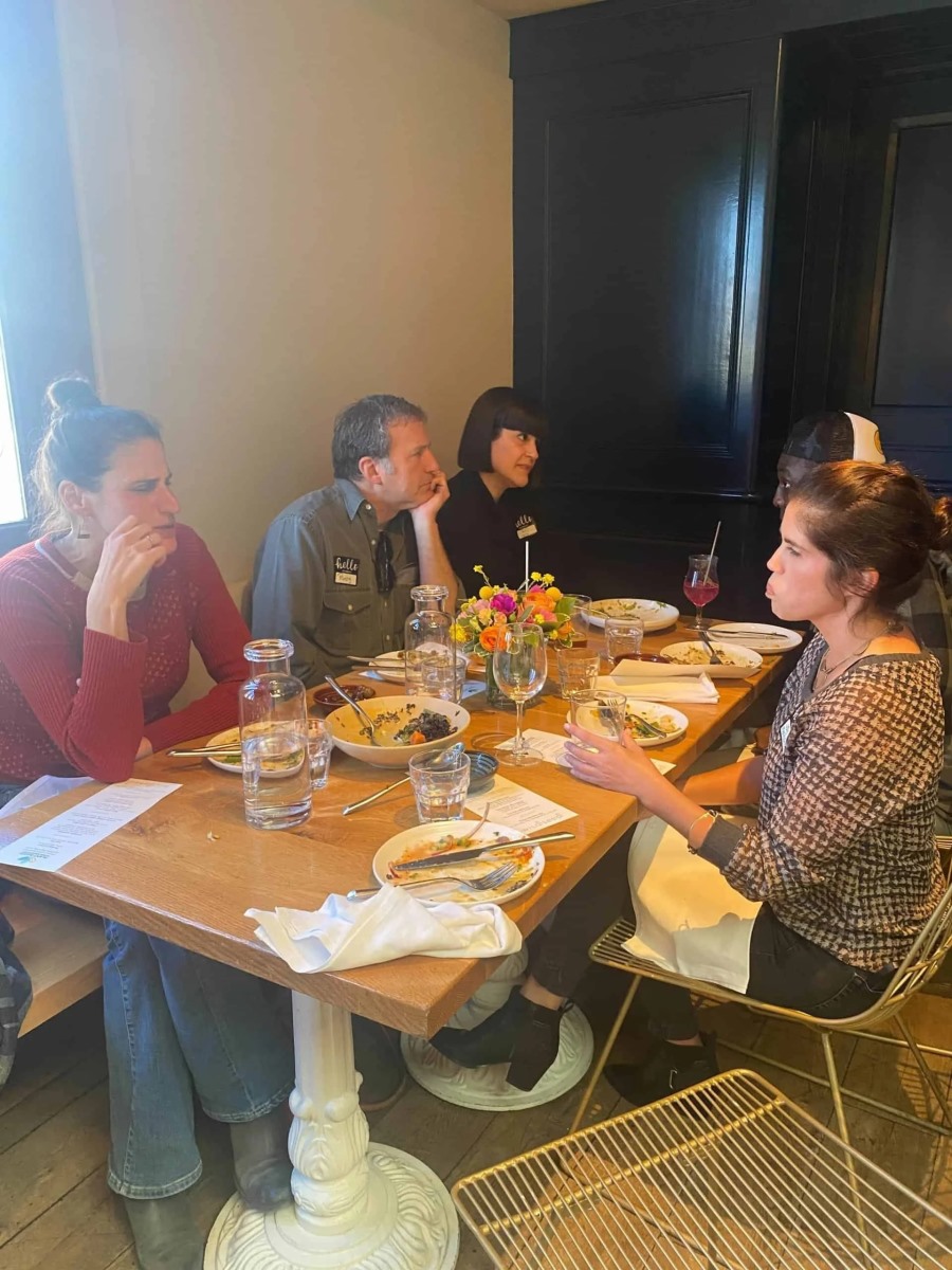 A group of five people sit together along a rustic wooden dining table in a restaurant, talking and gesturing over shared plates of food and a central bouquet of flowers.