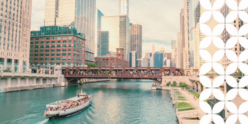 A view looking down the Chicago River lined with towering downtown skyscrapers. An architecture tour boat cruises along the water toward a brown steel bascule bridge spanning the river. A white geometric pattern frames the right edge of the image.