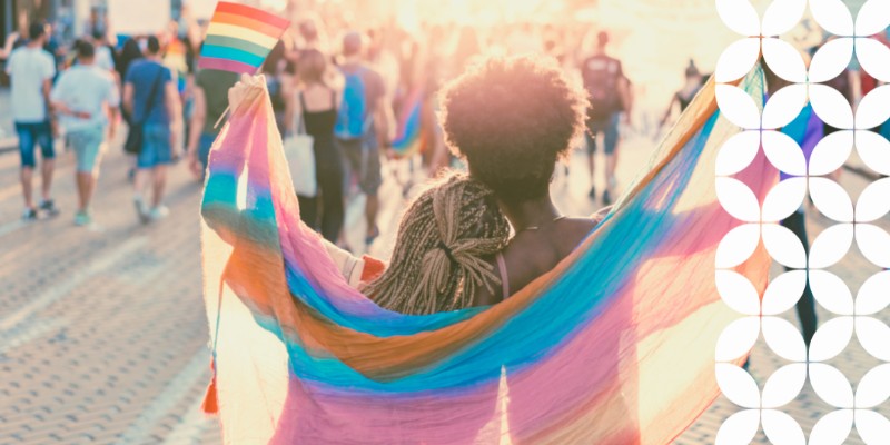 A view from behind of two people embracing at a crowded street festival during a sunny day. They are draped in a large, translucent rainbow Pride flag, with one person holding up a smaller rainbow hand flag. A white geometric pattern runs down the right border of the image.
