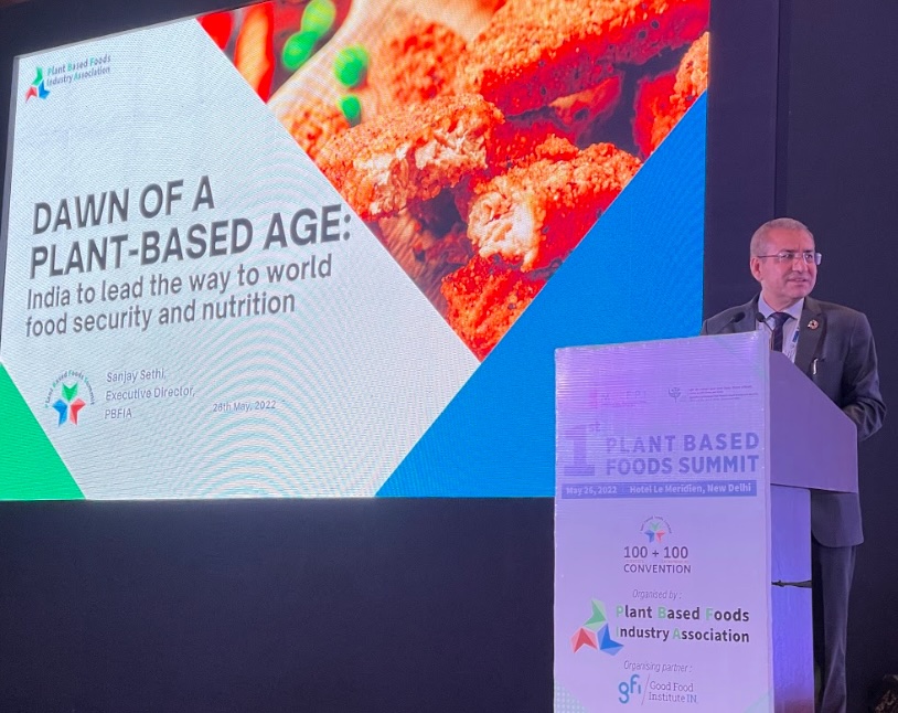 A man in a suit stands at a podium presenting at the 1st Plant Based Foods Summit in New Delhi. A large screen behind him displays a presentation slide titled "DAWN OF A PLANT-BASED AGE: India to lead the way to world food security and nutrition" alongside an image of plant-based food.
