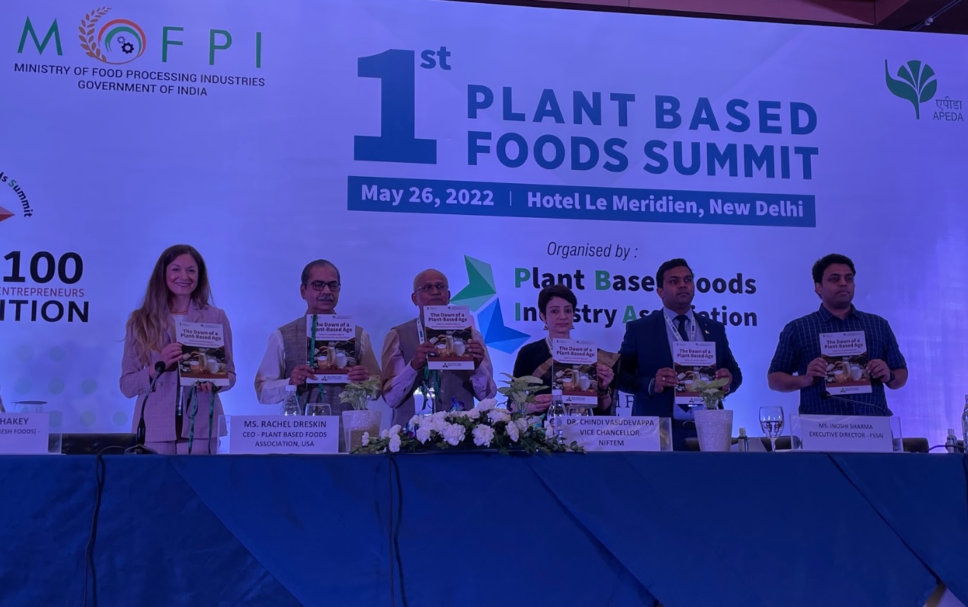 A lineup of six professionals standing behind a long, blue-draped table on a stage, each holding up a book titled "The Dawn of a Plant-Based Age." A large banner behind them marks the "1st Plant Based Foods Summit" hosted by the Plant Based Foods Industry Association.