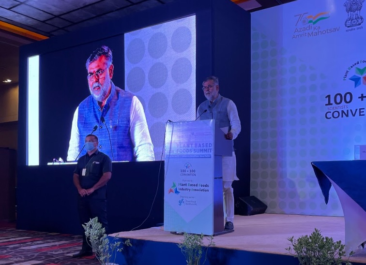 A man stands on a stage speaking at a podium during the Plant Based Foods Summit. A massive digital screen to his left mirrors his live presentation to the audience, while a masked event staff member stands nearby.