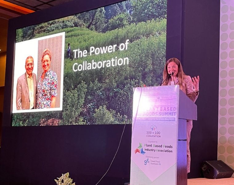 A woman gestures actively while speaking from a podium at the Plant Based Foods Summit. The backdrop screen reads "The Power of Collaboration" over a lush green hillside graphic, next to a photo of a man and a woman smiling.