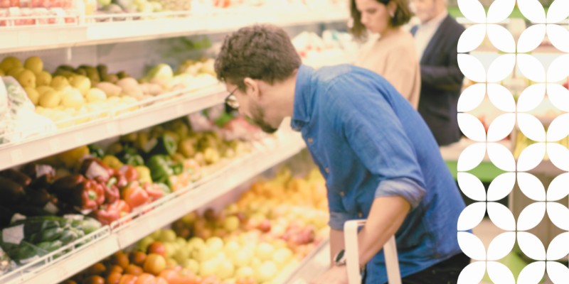 A man wearing a blue shirt leans forward to inspect fresh produce on a grocery store display shelf, with other shoppers visible but blurred in the background. A white geometric pattern frames the right edge of the image.