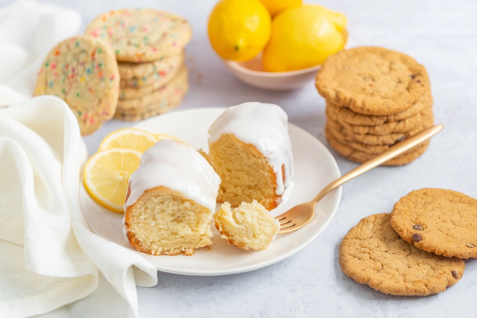 A close-up food flat lay of baked goods on a light gray surface. A white plate holds two slices of lemon bundt cake with white icing, next to real lemon slices and a gold fork. Surrounding the plate are stacks of colorful sprinkle sugar cookies and classic chocolate chip cookies.