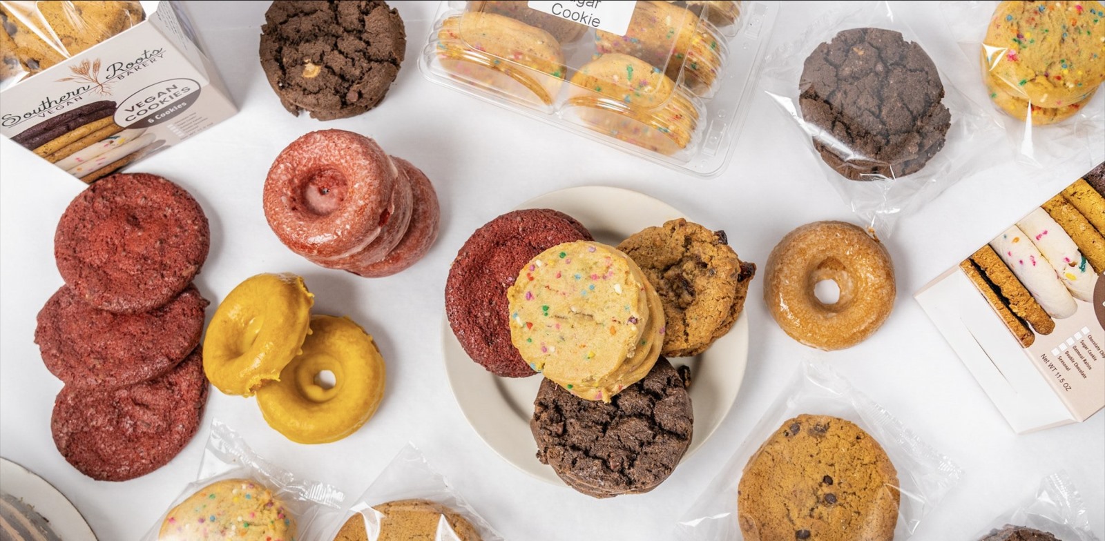 An overhead shot of a white surface scattered with various baked goods from Southern Roots Vegan Bakery, including loose and packaged glazed donuts, red velvet treats, chocolate cookies, and sprinkle-covered sugar cookies.