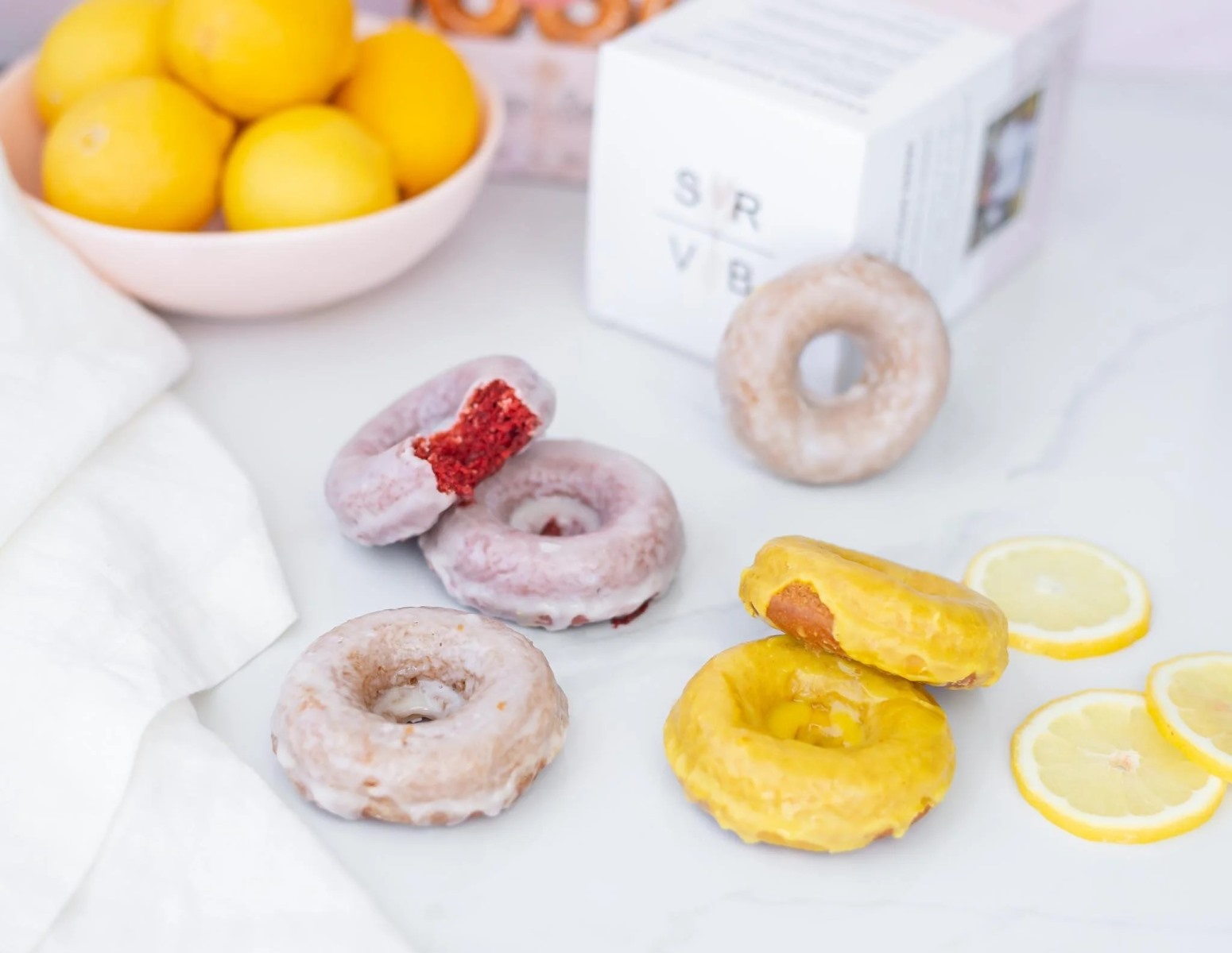 A bright tabletop arrangement of vegan donuts, showcasing iced red velvet donuts, yellow-glazed lemon donuts, and a classic glazed donut. A white box labeled "SRVB" and a bowl filled with whole fresh lemons are visible in the background.