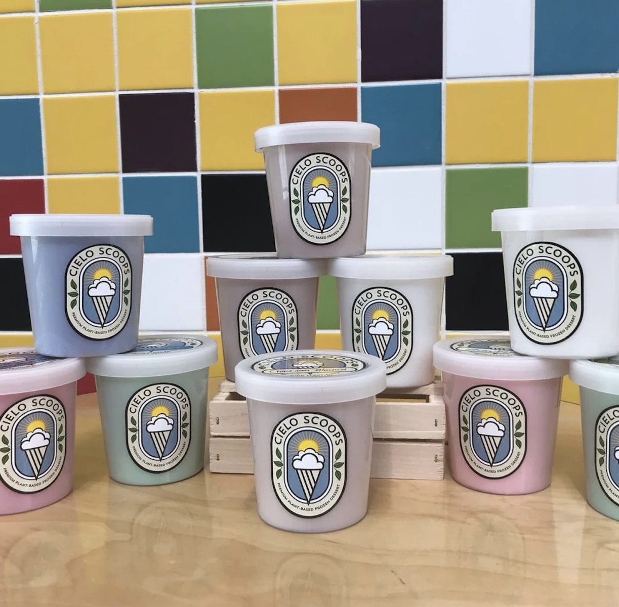 Several pastel-colored ice cream pints from "Cielo Scoops" stacked in a pyramid shape on a wooden table. The pints feature a logo with an ice cream cone in front of a sun, set against a background wall of multi-colored square tiles.