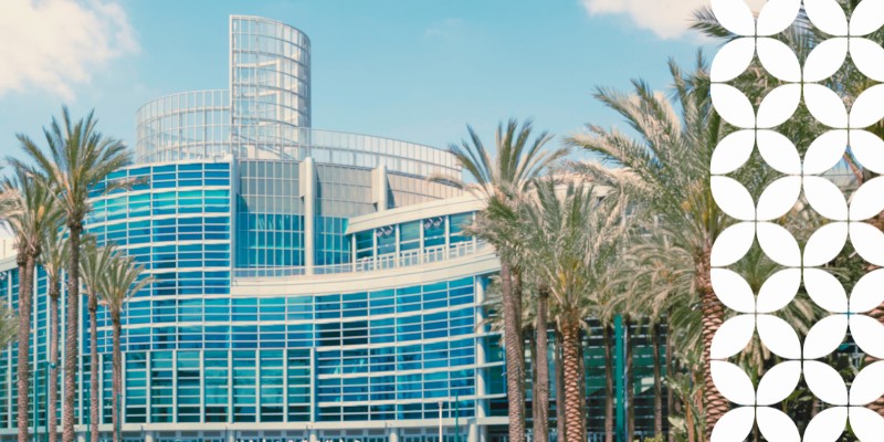 A wide exterior view of a large, modern convention center building featuring a curved glass facade and a glass rotunda on top. Tall palm trees line the front of the complex against a clear blue sky. A white geometric pattern runs down the right border of the image.