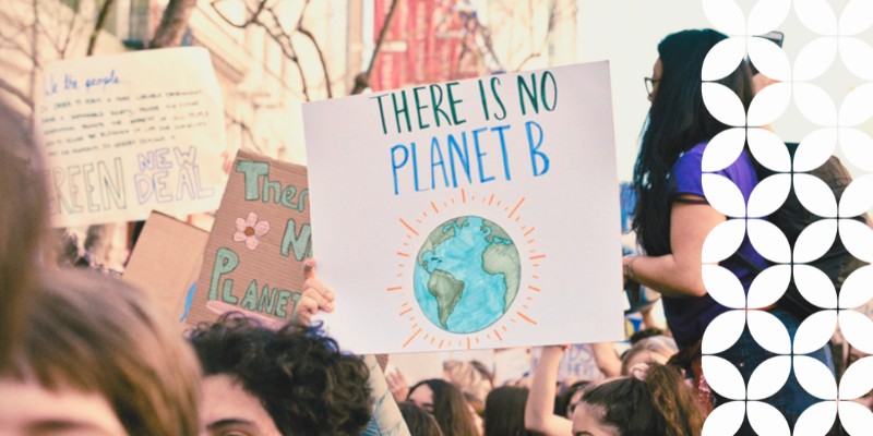 A close-up view of a crowd at a climate protest. An activist holds up a prominent white sign that reads "THERE IS NO PLANET B" above an illustration of the Earth. Other signs, including one that reads "GREEN NEW DEAL," are visible in the blurred background. A white geometric pattern runs down the right border of the image.