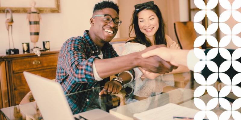A smiling young couple sits at a glass table during a meeting. The man, wearing glasses and a plaid shirt, reaches across the table to shake hands with someone out of frame, while a laptop and documents sit in the foreground. A white geometric pattern runs down the right border of the image.