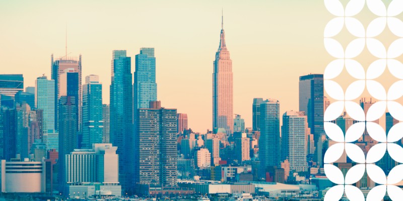 The midtown Manhattan skyline at dusk, showcasing the Empire State Building towering over the city buildings during a pink and golden sunset. A white geometric pattern runs down the right border of the image.