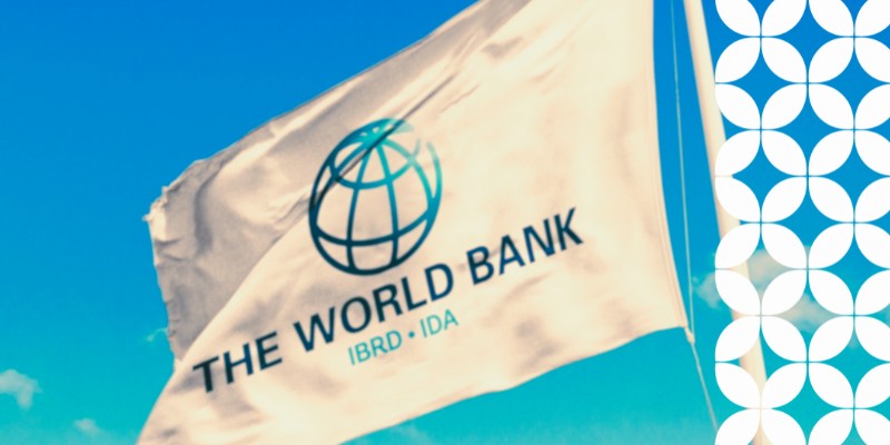 A close-up view of the white World Bank flag, featuring its blue globe logo and the words "THE WORLD BANK • IBRD • IDA," waving on a pole against a bright blue sky. A white geometric pattern runs down the right border of the image.