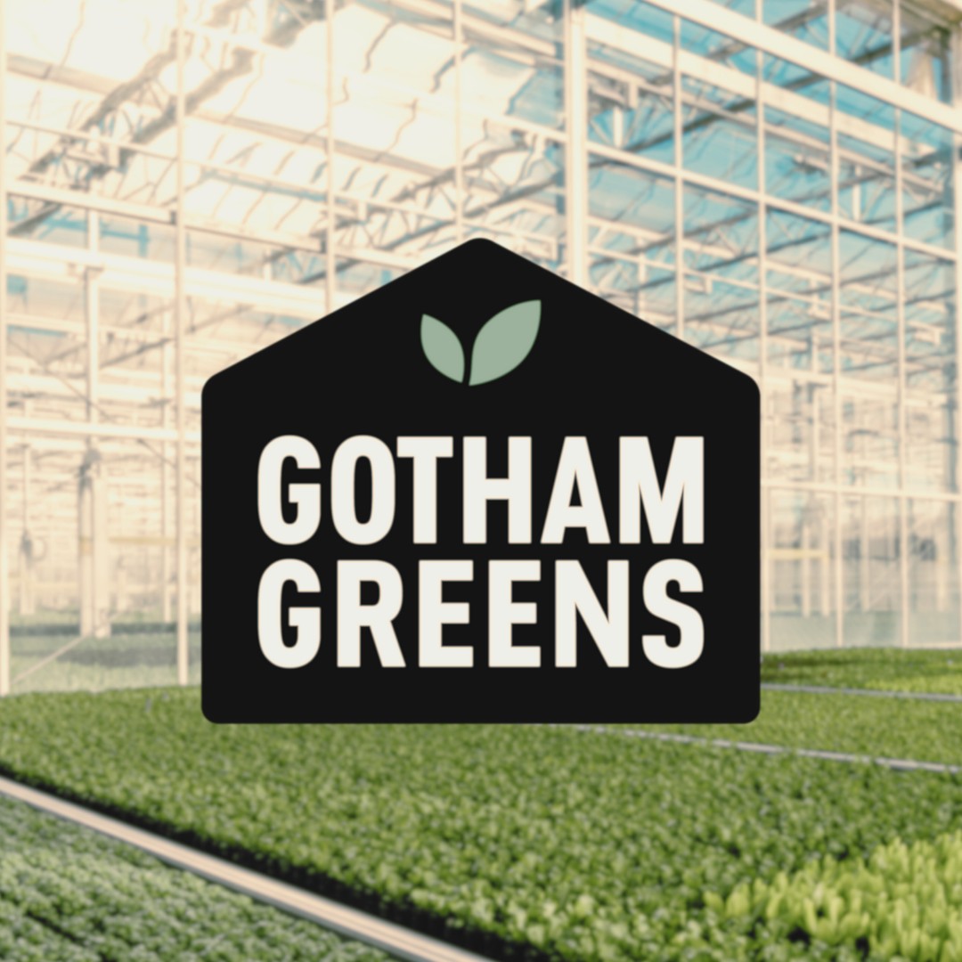 A large black house-shaped logo reading "GOTHAM GREENS" with a small two-leaf icon centered above the text, overlaid onto a background of structured greenhouse rows filled with plants.
