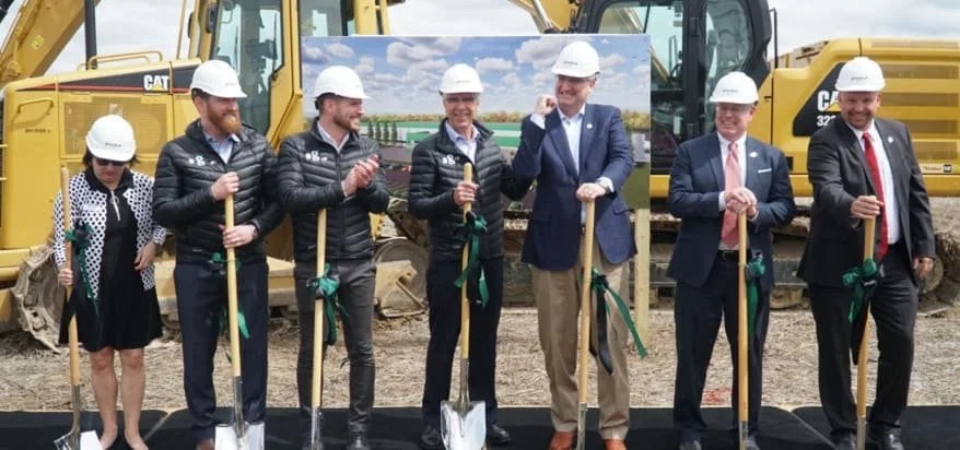 A group of seven dignitaries and corporate officials wearing white hard hats pose together in front of yellow heavy construction machinery during an outdoor groundbreaking ceremony. They hold ceremonial wooden shovels decorated with green ribbons on a black platform.